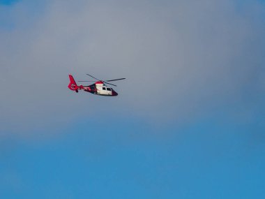 Mavi gökyüzünde kırmızı ve beyaz bir acil durum helikopteri uçuyor.