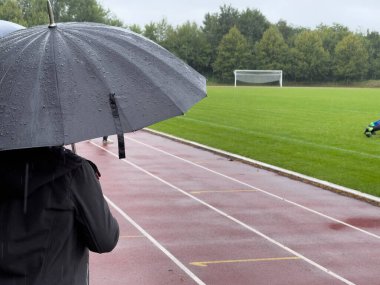 Bir insan yağmurda şemsiyeyle bir spor sahasının kenarında durur.