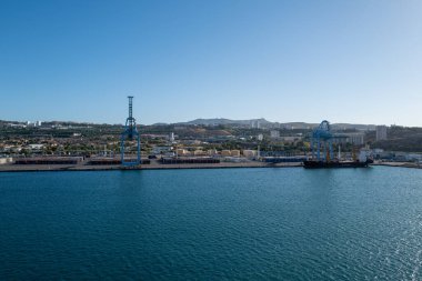Güneşli bir günde, derin mavi Akdeniz 'den geçen yolcu feribotu ardında beyaz bir iz bırakıyor. Arka planda, tepeli kıyı şeridi ve kıyı kasabası dağınık bulutlu açık mavi bir gökyüzünün altında görülebilir.. 