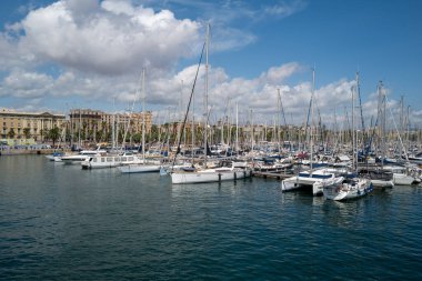 Akdeniz kıyı şehrinde güneşli bir günde yelkenli ve yatlarla dolu bir marina manzarası. Sakin mavi su ve berrak gökyüzü, kıyıya yanaşmış sayısız gemi direği ve yelkenli tekneleriyle güzel bir deniz havası yaratıyor. Perf