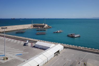 Güneşli bir günde, turkuaz mavi sularda yüzen su ürünleri ve balık çiftlikleriyle dolu bir limanın havadan görüntüsü. Görüntü dalgalı su duvarları, endüstriyel yapıları ve açık mavi gökyüzünün altındaki gemileri gösteren modern bir liman alanı gösteriyor. Perf