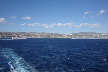 Güneşli bir günde, derin mavi Akdeniz 'den geçen yolcu feribotu ardında beyaz bir iz bırakıyor. Arka planda, tepeli kıyı şeridi ve kıyı kasabası dağınık bulutlu açık mavi bir gökyüzünün altında görülebilir.. 