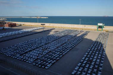 Limandaki büyük bir otomobil terminalinin hava görüntüsü, ihracat veya ithalatı bekleyen yeni araçları gösteriyor. Sahne, düzgün park edilmiş arabalar, nakliye konteynerleri ve rıhtımın yanındaki vinçlerle küresel otomotiv lojistiğinin ölçeğini yakalıyor.
