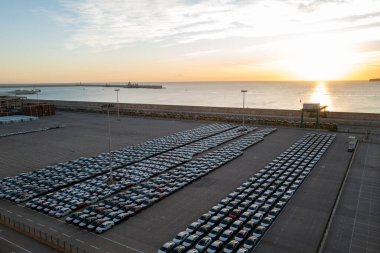 Limandaki büyük bir otomobil terminalinin hava görüntüsü, ihracat veya ithalatı bekleyen yeni araçları gösteriyor. Sahne, düzgün park edilmiş arabalar, nakliye konteynerleri ve rıhtımın yanındaki vinçlerle küresel otomotiv lojistiğinin ölçeğini yakalıyor.