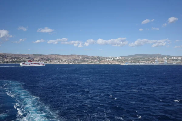 Güneşli bir günde, derin mavi Akdeniz 'den geçen yolcu feribotu ardında beyaz bir iz bırakıyor. Arka planda, tepeli kıyı şeridi ve kıyı kasabası dağınık bulutlu açık mavi bir gökyüzünün altında görülebilir.. 