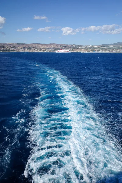 Güneşli bir günde, derin mavi Akdeniz 'den geçen yolcu feribotu ardında beyaz bir iz bırakıyor. Arka planda, tepeli kıyı şeridi ve kıyı kasabası dağınık bulutlu açık mavi bir gökyüzünün altında görülebilir. Görüntü beni yakalar.