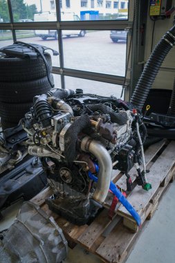 Bir otomotiv atölyesinin içindeki ahşap bir palete yerleştirilmiş modern bir araba motorunun detaylı bir yakın çekimi. Motorda görünür turbo şarj aleti, soğutucu borular ve kablo dizginleri, gelişmiş araç mühendisliği ve mekanik hassasiyet vurgulanıyor.