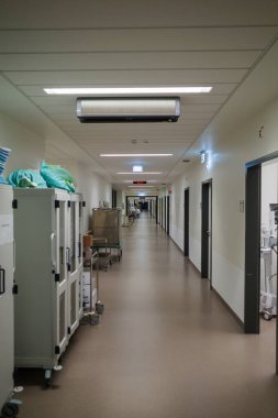 Hastane koridorunda, duvarlar boyunca tıbbi ekipman ve depolama dolapları olan temiz ve parlak bir ışık. Uzun ve steril koridorda birden fazla hasta odası, acil çıkış tabelaları ve modern ışıklandırma bulunuyor.