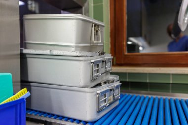 Stacked sterile metal instrument containers in a hospital sterilization room. The aluminum medical boxes, equipped with secure locking mechanisms and ventilation grids, sit on a blue roller conveyor, ready for cleaning, disinfection, or transport to 