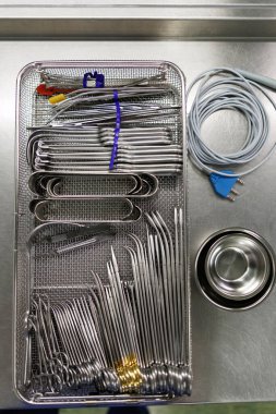 Hastanenin sterilizasyon odasındaki steril cerrahi aletlerle dolu bir tepsinin tepeden tırnağa görüntüsü. Paslanmaz çelik aletler arasında kıskaçlar, retraktörler, cımbızlar ve hassas cerrahi aletler var. Hepsi bir ağ sterilizati içinde düzenlenmiş.
