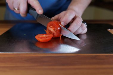 Mutfakta paslanmaz çelik bir kesme tahtasıyla taze bir domatesi dilimleyen birinin yakın çekimi. Eller ahşap bir tezgahta domates dilimleri keserken keskin bir mutfak bıçağını dikkatlice tutuyor. Sahne yemek hazırlama, evde yemek pişirme, o
