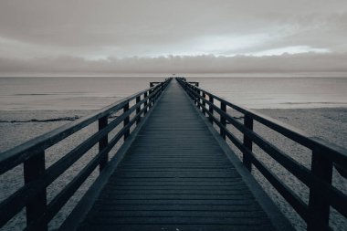 Minimalist deniz manzarası, bulutlu bir gökyüzünün altında sakin bir okyanusa doğru uzanan uzun bir ahşap iskeleden oluşuyor. Simetrik kompozisyon ve sessiz tonlar, yalnızlığı, yansımayı ve sonsuzluğu çağrıştıran, huzurlu ve karamsar bir atmosfer yaratıyor. Boş armut.