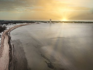 Gün batımında kıyı kentinin nefes kesen kış manzarası, ön planda karla kaplı tarlalar, sakin bir koy ve kıyı şeridinde modern bir gökdelen. Dramatik turuncu ve mor bulutlar gökyüzünde parlayarak çarpıcı bir c oluşturur.