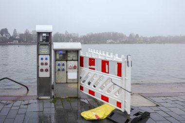 Almanya 'nın Eckernfrde kentindeki liman sahnesinde Baltık Denizi' nin yanındaki bir liman güvertesinde elektrik santralleri görülüyor. Geçici inşaat bariyerleri ve güvenlik ekipmanları ıslak kaldırımda duruyor.