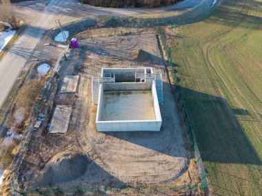 Beton bir su havzasının ya da kırsal alanda inşaat halindeki endüstriyel rezervlerin insansız hava aracı görüntüsü. Metal geçitli dikdörtgen beton yapı, altyapı gelişimini ön plana çıkaran tarım arazisi ve bir köy yolu ile çevrilidir.