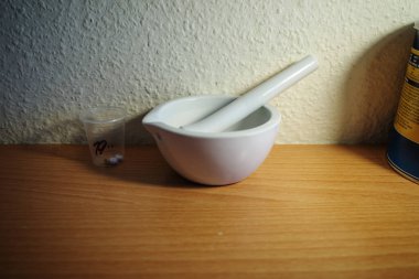 Minimalist natürmort fotoğraf beyaz seramik bir havan topu ve ahşap bir yüzey üzerinde haplar içeren saydam bir ilaç bardağının yanında tokmak gösteriyor. Olay yeri farmasötik hazırlık, ilaç, sağlık ve tıbbi tedavi öneriyor. Bu yüzden