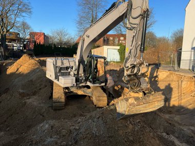 Geniş çaplı bir kazı alanı, açık mavi gökyüzünün altında kumlu topraklarda derin bir temel kazıyor. Bir inşaat işçisi ağır makinelerin yanında dururken toprak taşıma işi zemini inşa etmek için hazırlıyor.