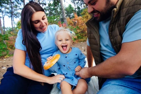 Liten flicka äter en bagel och skrattar glatt i naturen med föräldrar. — Stockfoto