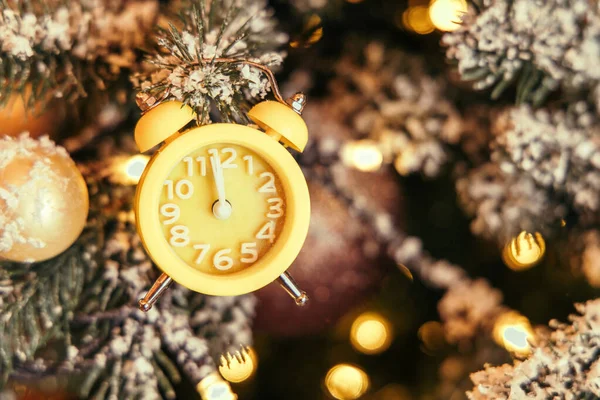 Close-up of a yellow clock showing the countdown to twelve o\'clock hangs on the Christmas tree next to the glowing golden lights on the background of New Year\'s decor. Merry Christmas.