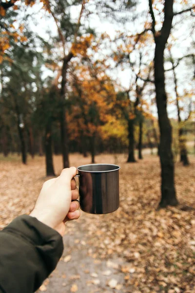 hand holding an iron cup with a hot drink on a background of autumn forest. vertical photo. mood of adventure and comfort.