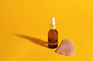 Sarı arka planda serumlu ya da yağlı kahverengi şişe ve sert gölgeli gua sha taşı. boşluktaki metin.