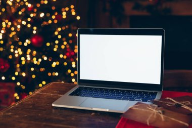 Beyaz ekran maketi olan dizüstü bilgisayar, karanlık ahşap bir masaya yerleştirilmiş, parlak ışıklandırılmış, bulanık bir Noel ağacının arka planına karşı.. 