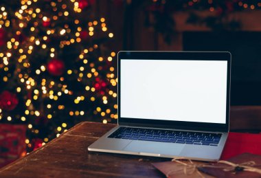 Beyaz ekran maketi olan dizüstü bilgisayar, karanlık ahşap bir masaya yerleştirilmiş, parlak ışıklandırılmış, bulanık bir Noel ağacının arka planına karşı.. 