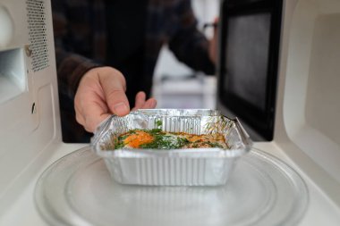 Adam ısıtmak için mikrodalga fırına çırpılmış yumurta ve otlarla folyo kabı koyuyor.