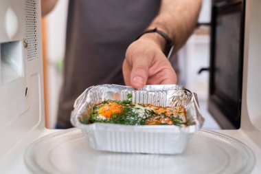 İnsan, mikrodalgaya çırpılmış yumurtalarla folyo tepsisi yerleştiriyor. Güvenli olmayan aygıt kullanımını vurguluyor.