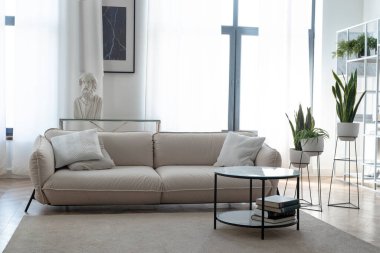 Modern, parlak bir oturma odası içinde bej bir kanepe, mermer bir sehpa, yeşil bitkiler ve klasik bir büst heykeli var. Minimalist apartman tasarımı