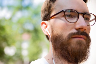 Sakallı ve gözlüklü genç bir adam, kulaklıkla müzik dinleyen bir hippi, yakın plan. Güneşli bir günde, açık havada müzikle rahatlamak..