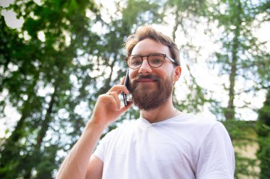 Güneşli bir günde dışarıda dikilirken beyaz tişörtlü, cep telefonuyla konuşan genç, sakallı bir adam. Cep telefonu, akıllı telefon kullanarak..