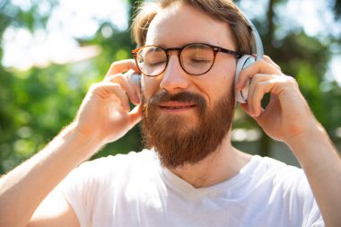 Genç sakallı, gülümseyen bir adam, gözleri kapalı bir hipster, kulaklıkla müzik dinliyor. Rahatlamak, müzikle meditasyon yapmak, güneşli bir günde dışarıda olmak.. 
