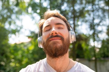 Genç, gülümseyen bir adam, gözleri kapalı bir hipster, kulaklıkla müzik dinliyor. Rahatlamak, müzikle meditasyon yapmak, güneşli bir günde dışarıda olmak.. 