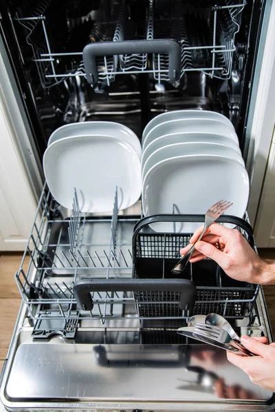Mãos masculinas carregando pratos e talheres em uma máquina de lavar louça aberta. Deveres domésticos com um aparelho de cozinha moderna. — Fotografia de Stock