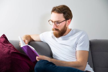 Young adult man reading a book, a magazine on a couch. Relax at home, in a bright modern interior.