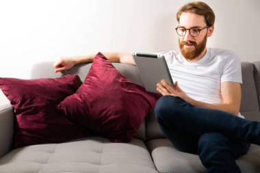 Young adult, bearded man using a tablet at home while sitting on a couch in a bright modern interior. Using an electronic, mobile device to browse the internet, to read newspaper.