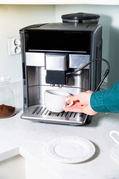 Making a coffee in a coffee machine, a coffee maker, at home, at office, in a bright kitchen. Male hand putting an empty cup for a coffee on a drip tray cover.