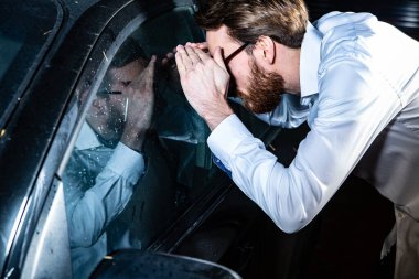 Young adult man looking through a side window into a car. Locked keys inside the car. A road accident, a car breakdown, emergency situation.