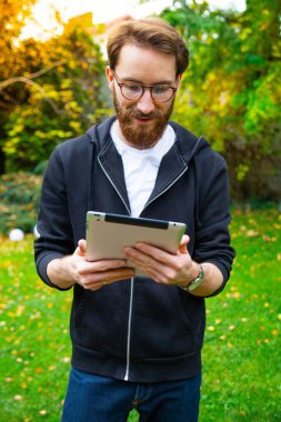 Genç yetişkin, sakallı bir adam güneşli bir günde açık havada, bahçede tablet kullanıyor. İnterneti gezmek, kitap okumak, gazete okumak için elektronik, mobil, taşınabilir bir cihaz kullanıyor..