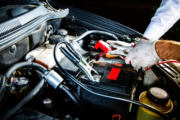 Male hands attaching clips to a car battery under a hood, at night, close-up. Jump starting a car engine with jumper cables on a car battery.