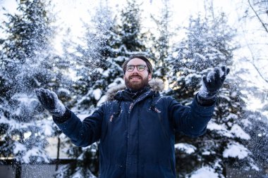 Kışlık giysiler içinde genç sakallı bir adam güneşli bir günde, kış manzarasında karlara serpilmişti. Kış eğlencesi, kartopu atma, açık hava eğlencesi..