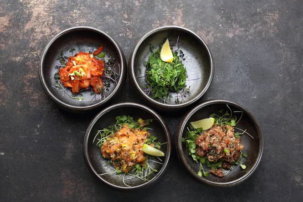 Composición de aperitivos: tártaro de atún, tártaro de salmón, kimchi, ensalada japonesa de algas marinas sobre un fondo pedregoso negro. — Foto de Stock