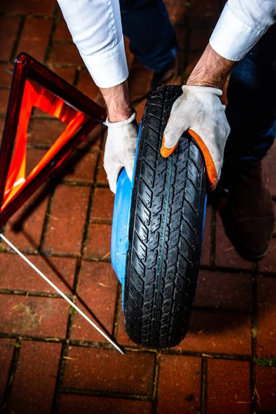 Vue verticale des mains mâles tenant une roue de secours, un triangle d'avertissement d'urgence en arrière-plan. Changer un pneu de voiture la nuit. — Photo