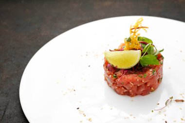 Zarif beyaz bir tabakta, siyah taşlı arka planda bir somon tartarı. Taze çiğ somon ve baharatlardan yapılmış mezeler, limon dilimiyle süslenmiş..