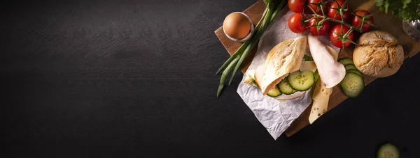 Ontbijt concept. Brede banner met sandwich met gevogelte worst, kaas, komkommer, een zacht gekookt ei, brood, groenten op een snijplank op zwarte stenige werkblad, achtergrond, met een copyspace. — Stockfoto