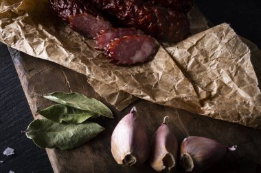 Tütsülenmiş sosis, yiyecek ambalajı kağıdı, sarımsak ve defne yapraklarıyla yatay, sade bir kompozisyon siyah taşlı bir tezgahın üzerinde eski usul tahta kesme tahtası. Polonyalı, ev yapımı sosis..