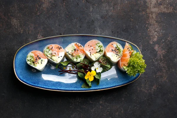 Bovenaanzicht van 6-delige sushi set met zalm, komkommer, filenkaas, in rijstpapier, versierd met eetbare bloem, op een blauwe plaat, op zwarte stenige ondergrond. Oosterse broodjes met zalm en groenten. — Stockfoto