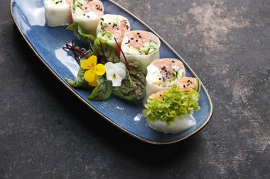 Somon, salatalık, Philadelphia peyniri ve pirinç kağıdıyla kaplanmış, yenilebilir çiçeklerle süslenmiş, mavi bir tabakta, siyah taşlı arka planda 6 parça suşi. Somon ve sebzeli Doğu dürümleri..