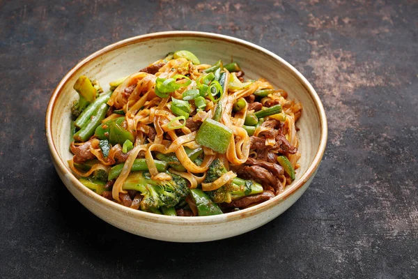 El primer plano de los tallarines orientales en la escudilla, sobre el fondo negro pedregoso. Carne frita en un wok con fideos de arroz, judías verdes, calabacín, pak choi y pimientos verdes en salsa picante de soja y ostras. Plato de cousine tailandés — Foto de Stock